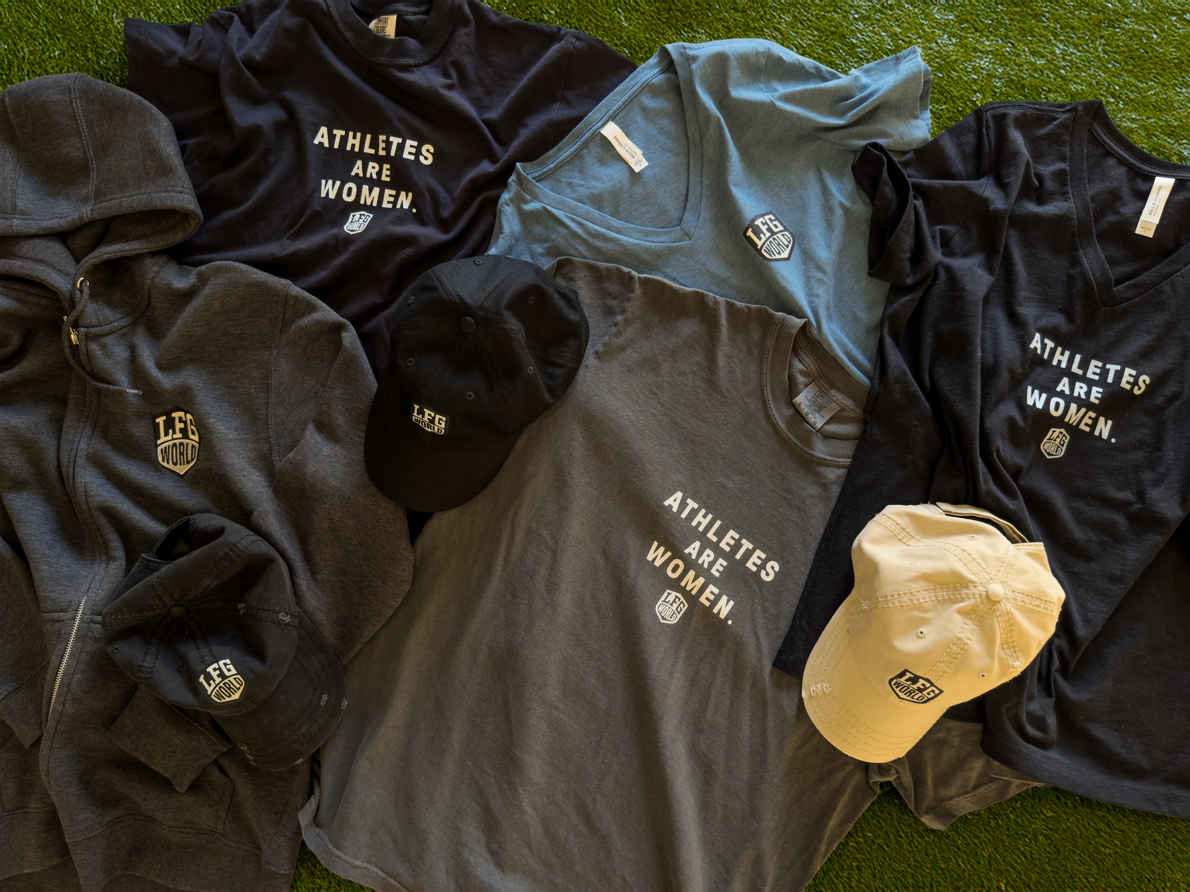 Collection of sports-themed apparel including t-shirts, hoodies, and caps on a grassy surface.